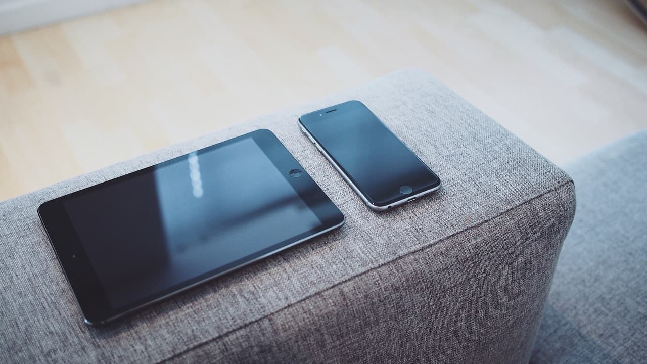 A tablet and a smartphone resting on the arm of a grey sofa